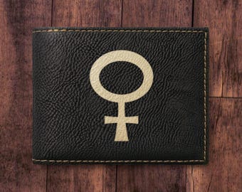 May include: A black bi-fold wallet with a textured surface featuring a centered, light-colored Venus symbol. The wallet is rectangular in shape with visible stitching along the edges, providing a clear contrast between the dark material and the icon.
