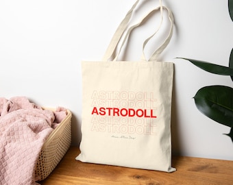 May include: A cream-colored canvas tote bag featuring the text ASTRODOLL repeated five times in a stacked design. The middle line is printed in bold red, while the others are in a thin red outline. Below the text is the phrase Have A Nice Day.
