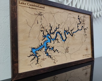 May include: A rectangular wooden wall map of Lake Cumberland, Kentucky, featuring a dark brown frame and a light wood surface. The lake is depicted with a bright blue inlay, surrounded by engraved black road lines and text labels.
