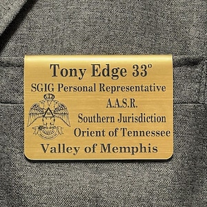 May include: A gold-coloured metal money clip with engraved text. The text includes a name, title, and organisational details, along with a symbol of an eagle with outstretched wings. The clip is rectangular with rounded corners.