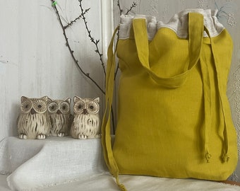 May include: A yellow linen tote bag with a white linen lining and drawstring closure. The bag is sitting on a white linen surface with three ceramic owl figurines.