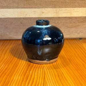 Könnte beinhalten: Eine kleine, runde, schwarze Keramikvase mit braunem Rand. Die Vase hat eine glänzende Oberfläche und eine dezente weiße Glasur.