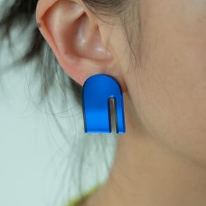 Puede incluir: Primer plano de un pendiente azul con un diseño en forma de U. El pendiente es de color azul vibrante y tiene un acabado brillante. El pendiente se lleva en la oreja de una persona. El fondo está borroso.