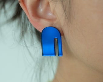 May include: A close-up of a blue earring with a U-shaped design. The earring is a vibrant blue color and has a glossy finish. The earring is worn on the ear of a person. The background is blurred.