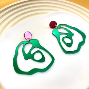 以下が含まれることがあります： 緑とピンクの抽象的な形のイヤリングのペア。イヤリングはアクリル製で、光沢のある仕上げになっています。