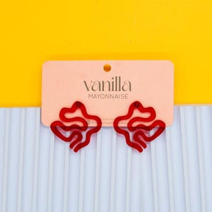 May include: Red acrylic earrings in a unique abstract shape. The earrings are on a pink card with the text "vanilla MAYONNAISE".