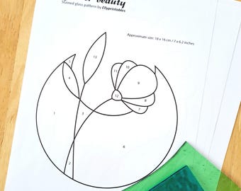May include: A white paper with a black line drawing of a flower, labeled "Flower beauty" and "Stained glass pattern by Ellyprintables." Approximate size is 18 x 16 cm. Two pieces of green glass are in the lower right corner.