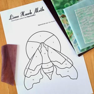 May include: A stained glass pattern labelled "Lime Hawk Moth" by Ellyprintables, approximately 16 x 16 cm in size. The design shows a moth outline with numbered sections, alongside colourful glass samples. A piece of purple glass is also present.