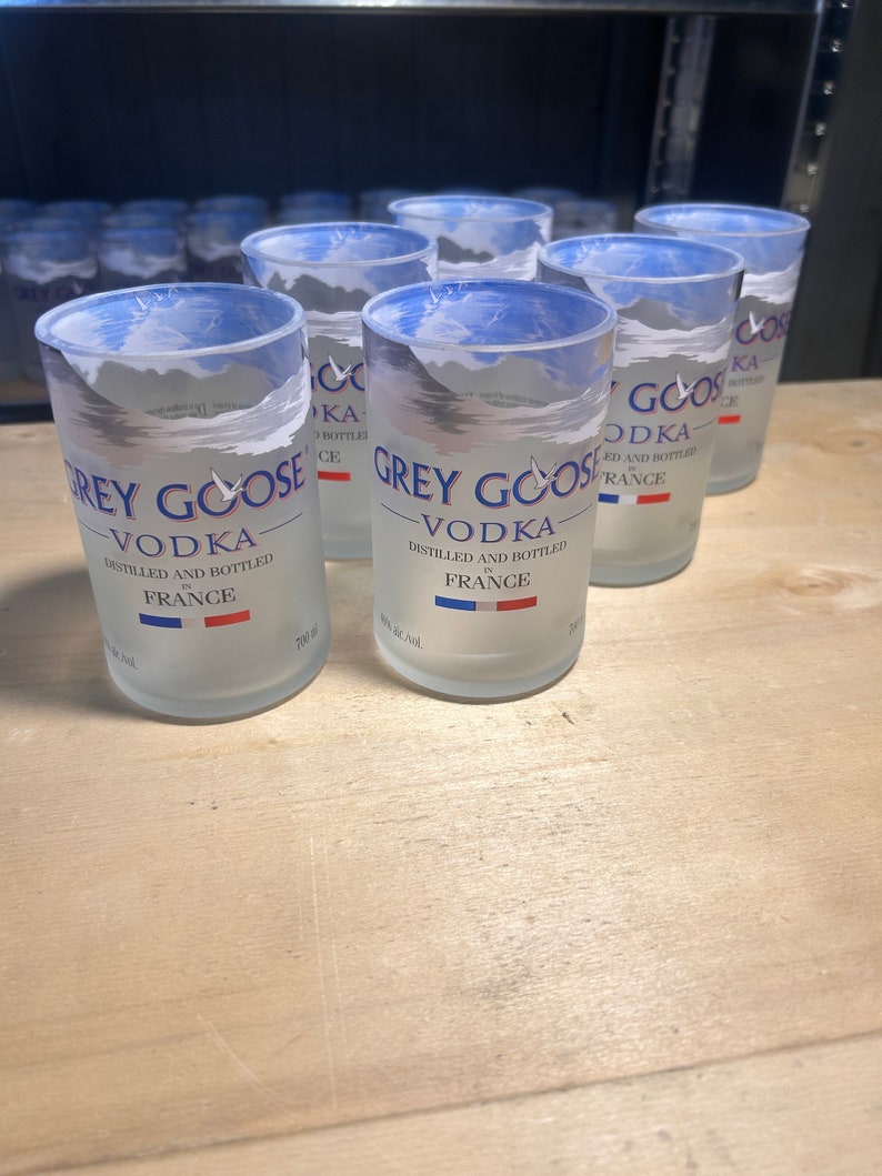 Pu&ograve; includere: Un gruppo di sei bottiglie di vodka Grey Goose in vetro trasparente con finitura opaca. Le bottiglie sono disposte in fila su una superficie di legno chiaro. Le bottiglie sono etichettate con il logo Grey Goose Vodka e le parole "Distillato e imbottigliato in Francia".