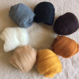 May include: A collection of eight balls of wool in various colors, including white, gray, black, brown, beige, and yellow. The balls are arranged in a circle on a light brown fabric background.