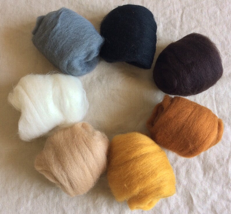 May include: A collection of eight balls of wool in various colors, including white, gray, black, brown, beige, and yellow. The balls are arranged in a circle on a light brown fabric background.