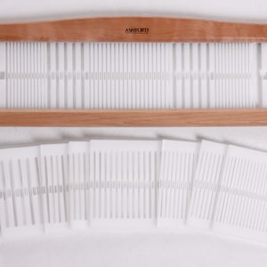 May include: A wooden Ashford Rigid Heddle Loom with a set of 10 white plastic heddle cards. The loom is made of light brown wood and has a curved top. The heddle cards are rectangular and have a series of holes in them.