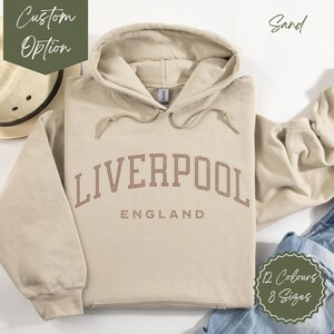 Può includere: Felpa con cappuccio color sabbia con il testo "LIVERPOOL ENGLAND" in lettere marroni.