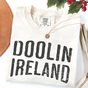 May include: Cream-colored t-shirt with the text "DOOLIN IRELAND" in distressed black lettering. A gold necklace with a pendant rests on the shirt. A brown hat and sprigs of red berries and green leaves are in the background.