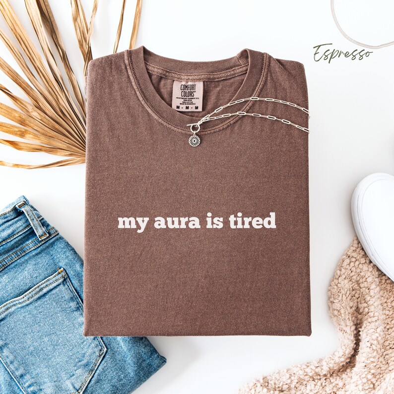 Flat lay Comfort Colors T Shirt in espresso with ‘my aura is tired’ written in bold white lower case text