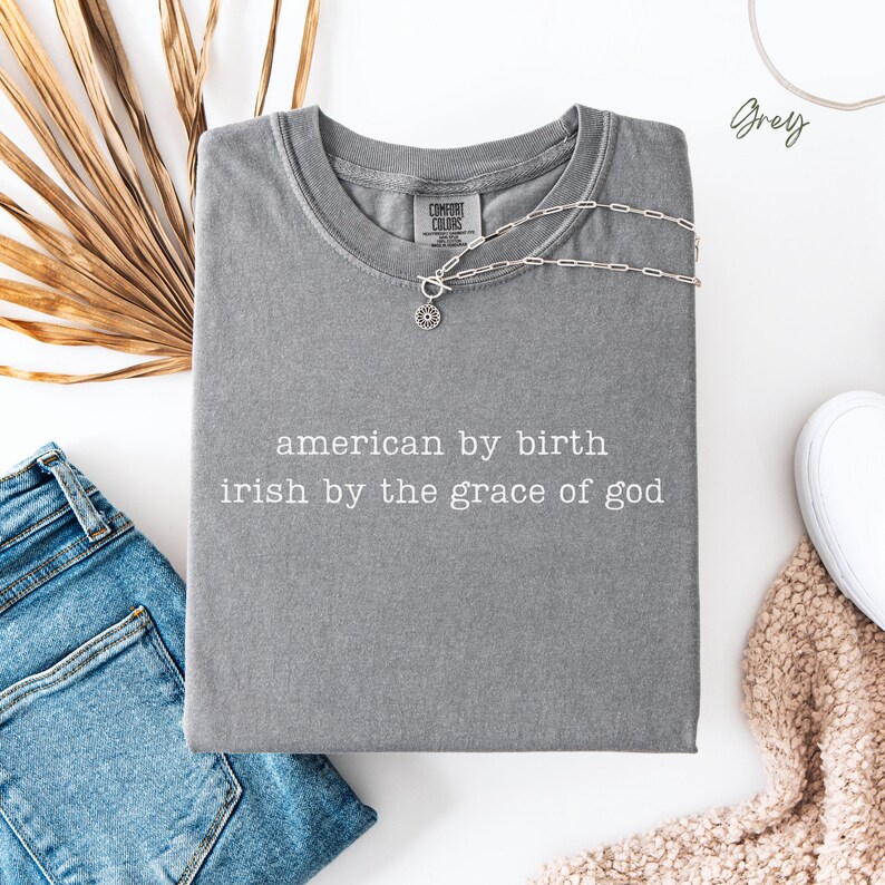 Flat lay of a folded grey Comfort Colors T-shirt featuring the quote “american by birth, irish by the grace of god” in lowercase typewriter text. Styled with denim jeans to the left, dried leaves at the back, a white sneaker and cream blanket on the bottom left, “grey” marked top right.