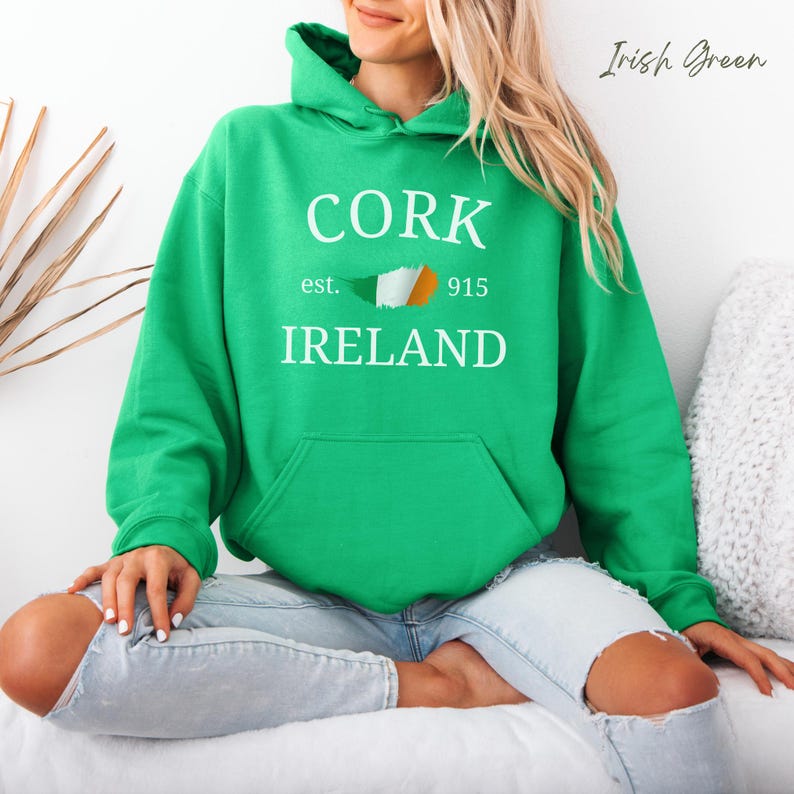 Woman wearing Irish Green hoodie Cork Ireland est 915 and Irish Flag. Sits casually cross legged wearing denim jeans, bare feet against comfy cushions & a wooden light colored backdrop. 
Text is clean and bold. Aesthetic is relaxed and casual.