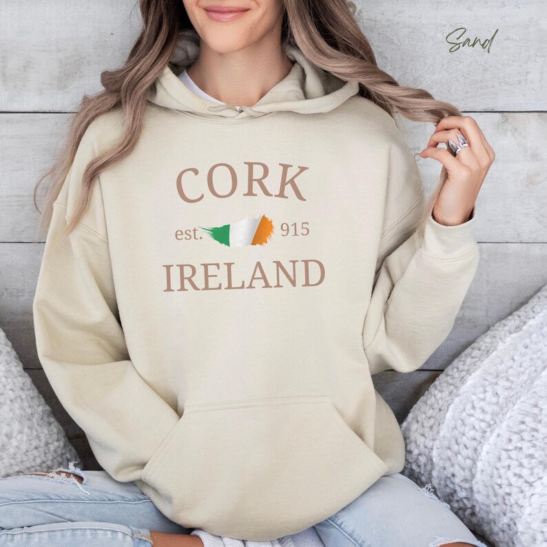 Woman wearing sand beige hoodie Cork Ireland est 915 and Irish Flag. Sits casually cross legged, denim jeans, cozy socks against comfy cushions & a wooden light colored backdrop. 
Text is clean and bold. Aesthetic is relaxed and casual.