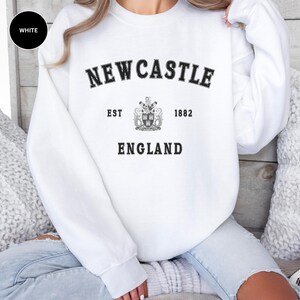 Woman wears white sweatshirt with Newcastle  England, EST 1882 and the Newcastle coat of arms in White distressed text. Also in Black, Dark Heather, Forest Green, forest green, Sand, Sport Grey and navy. Sizes from Small to XXL.