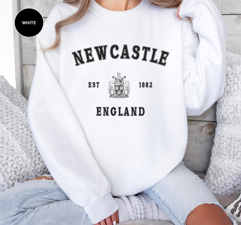 Woman wears white sweatshirt with Newcastle  England, EST 1882 and the Newcastle coat of arms in White distressed text. Also in Black, Dark Heather, Forest Green, forest green, Sand, Sport Grey and navy. Sizes from Small to XXL.