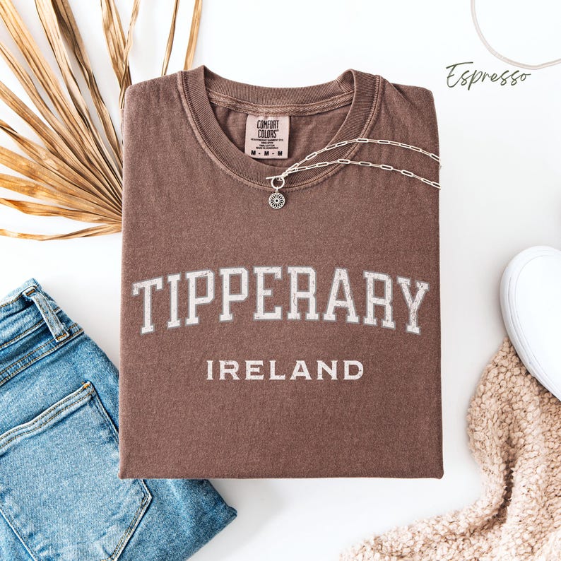 Flat Lay folded comfort colors® T shirt in Espresso Brown with Tipperary, Ireland in college style distressed look lettering. The shirt is in the centre of the picture with a pair of blue jeans to the left, some brown dried foliage sitting to the back left of the image.