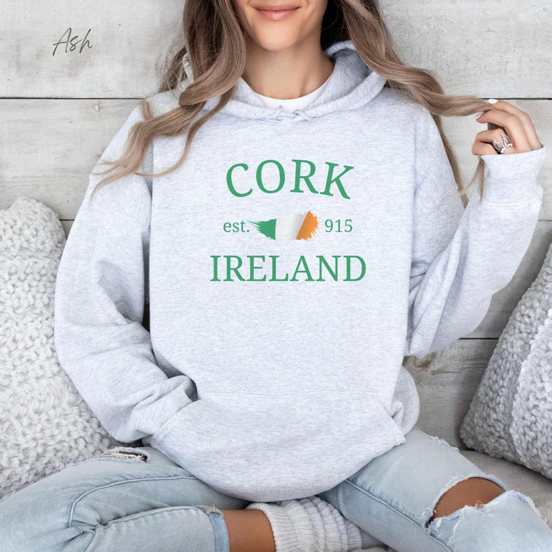 Woman wearing ash grey hoodie Cork Ireland est 915 and Irish Flag. Sits casually cross legged, denim jeans, cozy socks against comfy cushions & a wooden light colored backdrop. 
Text is clean and bold. Aesthetic is relaxed and casual.