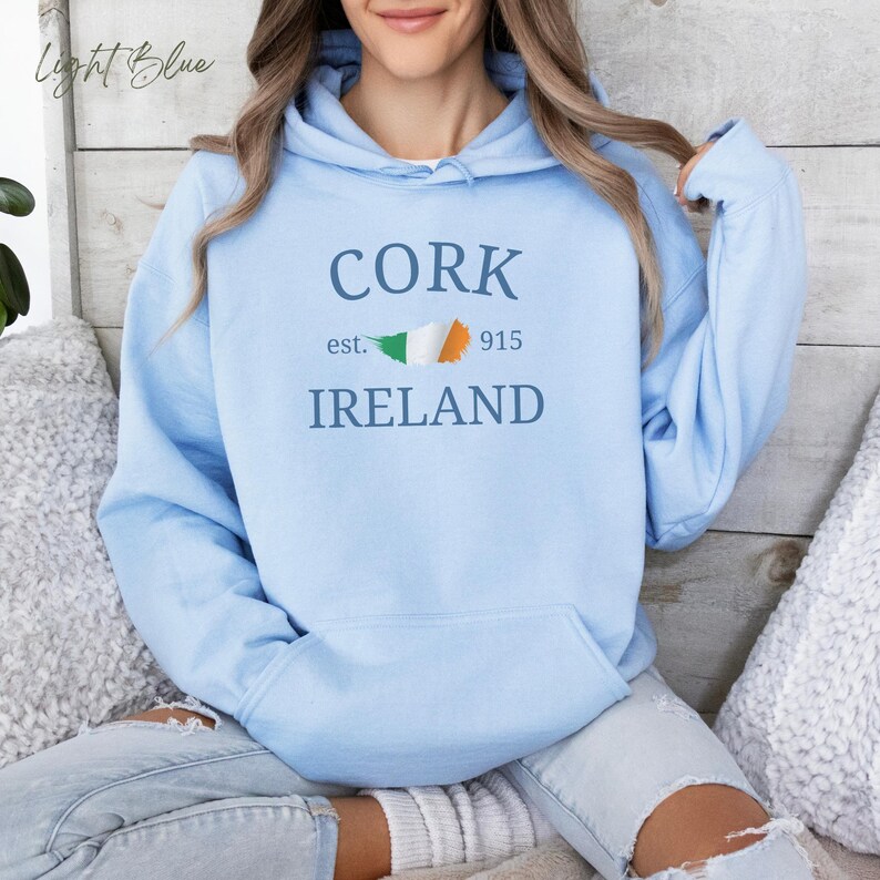 Woman wearing light blue hoodie Cork Ireland est 915 and Irish Flag. Sits casually cross legged, denim jeans, cozy socks against comfy cushions & a wooden light colored backdrop. 
Text is clean and bold. Aesthetic is relaxed and casual.