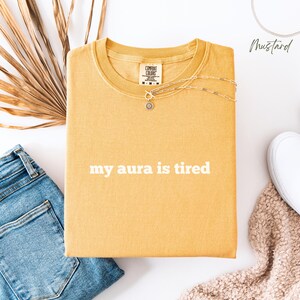 Flat lay Comfort Colors T Shirt in mustard with ‘my aura is tired’ written in bold white lower case text