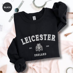 Op de afbeelding: Zwart sweatshirt met ronde hals met het woord "LEICESTER" in wit, gebogen over de borst. Onder de tekst staat een wapen en het woord "ENGLAND". Het sweatshirt heeft ook de tekst "EST. 1919".