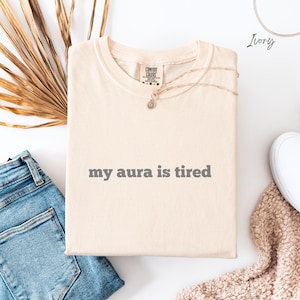Flat lay Comfort Colors T Shirt in ivory with ‘my aura is tired’ written in bold grey lower case text