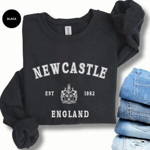 Lay flat black sweatshirt with Newcastle  England, EST 1882 and the Newcastle coat of arms in White distressed text. Also in royal, Dark Heather, Forest Green, Navy, Sand, Sport Grey and White. Sizes from Small to XXL.