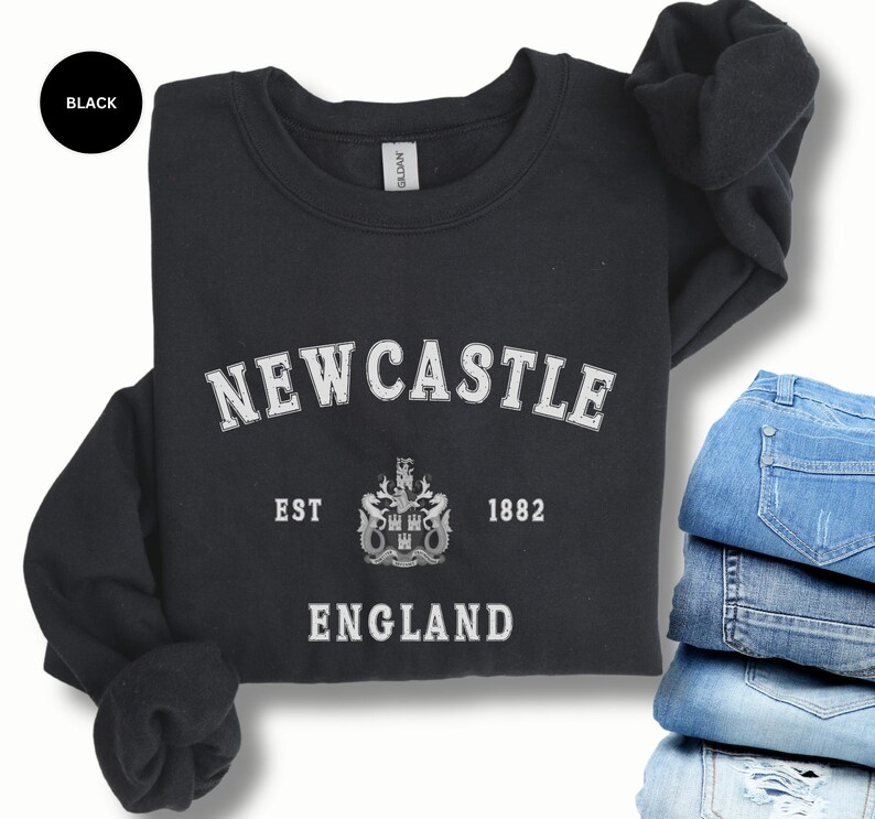 Lay flat black sweatshirt with Newcastle  England, EST 1882 and the Newcastle coat of arms in White distressed text. Also in royal, Dark Heather, Forest Green, Navy, Sand, Sport Grey and White. Sizes from Small to XXL.