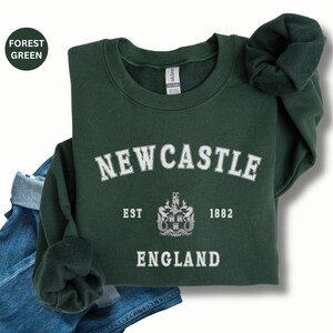 Lay flat forest green sweatshirt with Newcastle  England, EST 1882 and the Newcastle coat of arms in White distressed text. Also in royal, Black, Dark Heather, Forest Green, Navy, Sand, Sport Grey and White. Sizes from Small to XXL.