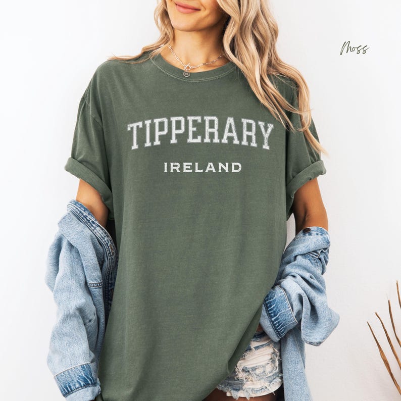 Tall blond woman in cut off denim shorts wearing comfort colors Moss Green  T shirt with Tipperary, Ireland in college style distressed look lettering. Shirt is worn oversize with an oversize blue denim jacket worn off the shoulder to give a minimalistic stylish look.