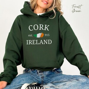 Puede incluir: Sudadera con capucha verde bosque con texto blanco que dice "CORK est. 915 IRLANDA" y un peque&ntilde;o gr&aacute;fico de bandera irlandesa.