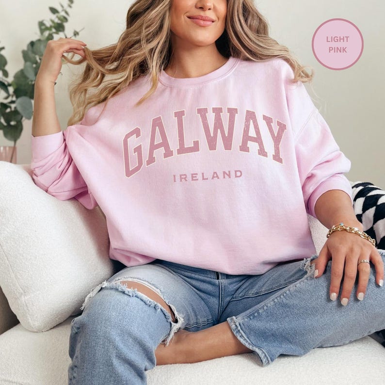 Woman wears light pink sweatshirt with Galway, Ireland in a duty rose pink vintage style text. Option to customise.