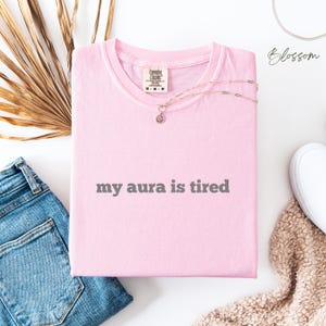 Flat lay Comfort Colors T Shirt in blossom pink with ‘my aura is tired’ written in bold grey lower case text