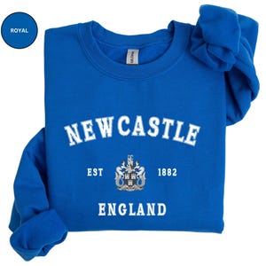 Lay flat royal blue sweatshirt with Newcastle  England, EST 1882 and the Newcastle coat of arms in White distressed text. Also in Black, Dark Heather, Forest Green, Navy, Sand, Sport Grey and White. Sizes from Small to XXL.