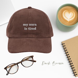May include: A dark brown corduroy baseball cap with white embroidered text that reads "my aura is tired".