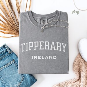 Flat Lay folded comfort colors® T shirt in Grey with Tipperary, Ireland in college style distressed look lettering. The shirt is in the centre of the picture with a pair of blue jeans to the left, some brown dried foliage sitting to the back left of the image.