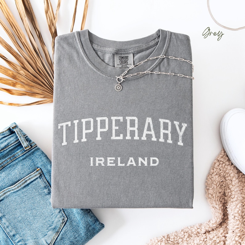 Flat Lay folded comfort colors® T shirt in Grey with Tipperary, Ireland in college style distressed look lettering. The shirt is in the centre of the picture with a pair of blue jeans to the left, some brown dried foliage sitting to the back left of the image.
