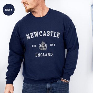 Man wears navy sweatshirt with Newcastle  England, EST 1882 and the Newcastle coat of arms in White distressed text. Also in Black, Dark Heather, Forest Green, forest green, Sand, Sport Grey and White. Sizes from Small to XXL.