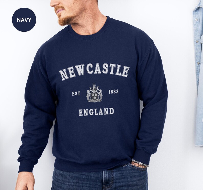 Man wears navy sweatshirt with Newcastle  England, EST 1882 and the Newcastle coat of arms in White distressed text. Also in Black, Dark Heather, Forest Green, forest green, Sand, Sport Grey and White. Sizes from Small to XXL.