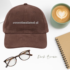 May include: A dark brown corduroy baseball cap with white embroidered text that reads "overstimulated af".