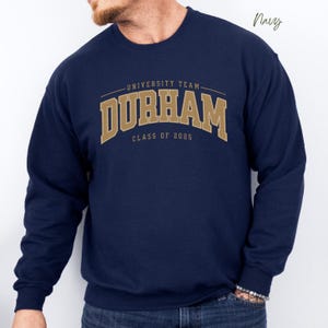 Pode incluir: Camisola azul marinho com letras douradas que dizem "University Team Durham Class of 2025".