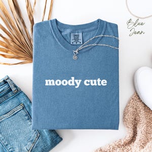 Könnte beinhalten: Ein blaues Jeans-T-Shirt mit dem Text "moody cute" in weißen Buchstaben. Das Shirt liegt flach auf einer weißen Oberfläche mit einer Jeans und einem weißen Schuh im Hintergrund.