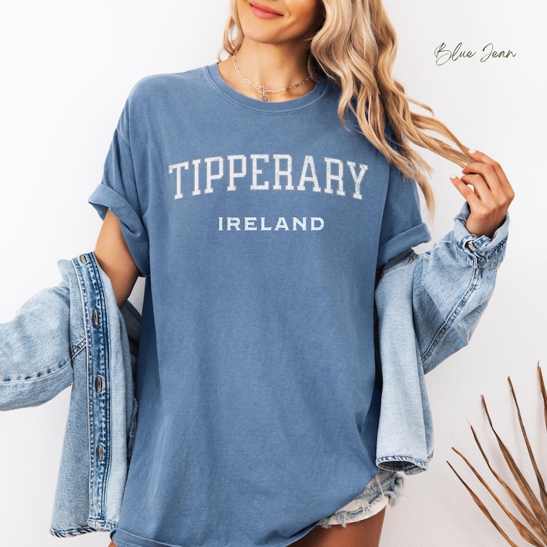 Tall blond woman in cut off denim shorts wearing comfort colors Blue Jean  T shirt with Tipperary, Ireland in college style distressed look lettering. Shirt is worn oversize with an oversize blue denim jacket worn off the shoulder to give a minimalistic stylish look.