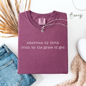 May include: A maroon t-shirt with the text "american by birth irish by the grace of god" printed in white. The shirt is made by Comfort Colors.