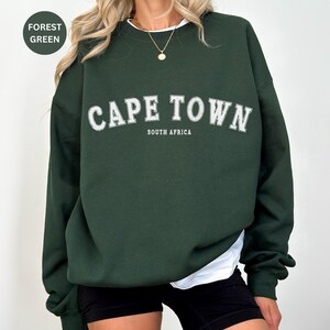 May include: A forest green sweatshirt with the text "CAPE TOWN" in white letters with a white outline. The text "SOUTH AFRICA" is printed in smaller white letters below the main text.