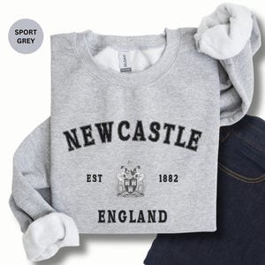 Lay flat sport grey sweatshirt with Newcastle  England, EST 1882 and the Newcastle coat of arms in White distressed text. Also in Black, Dark Heather, Forest Green, Navy, Sand, royal blue and White. Sizes from Small to XXL.